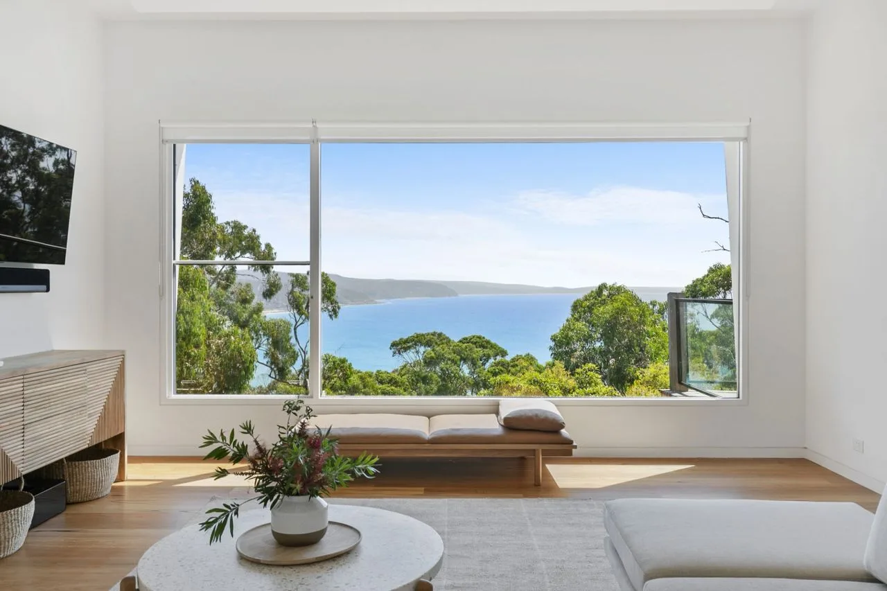 A bright living room opens onto a wide window overlooking trees and the water.