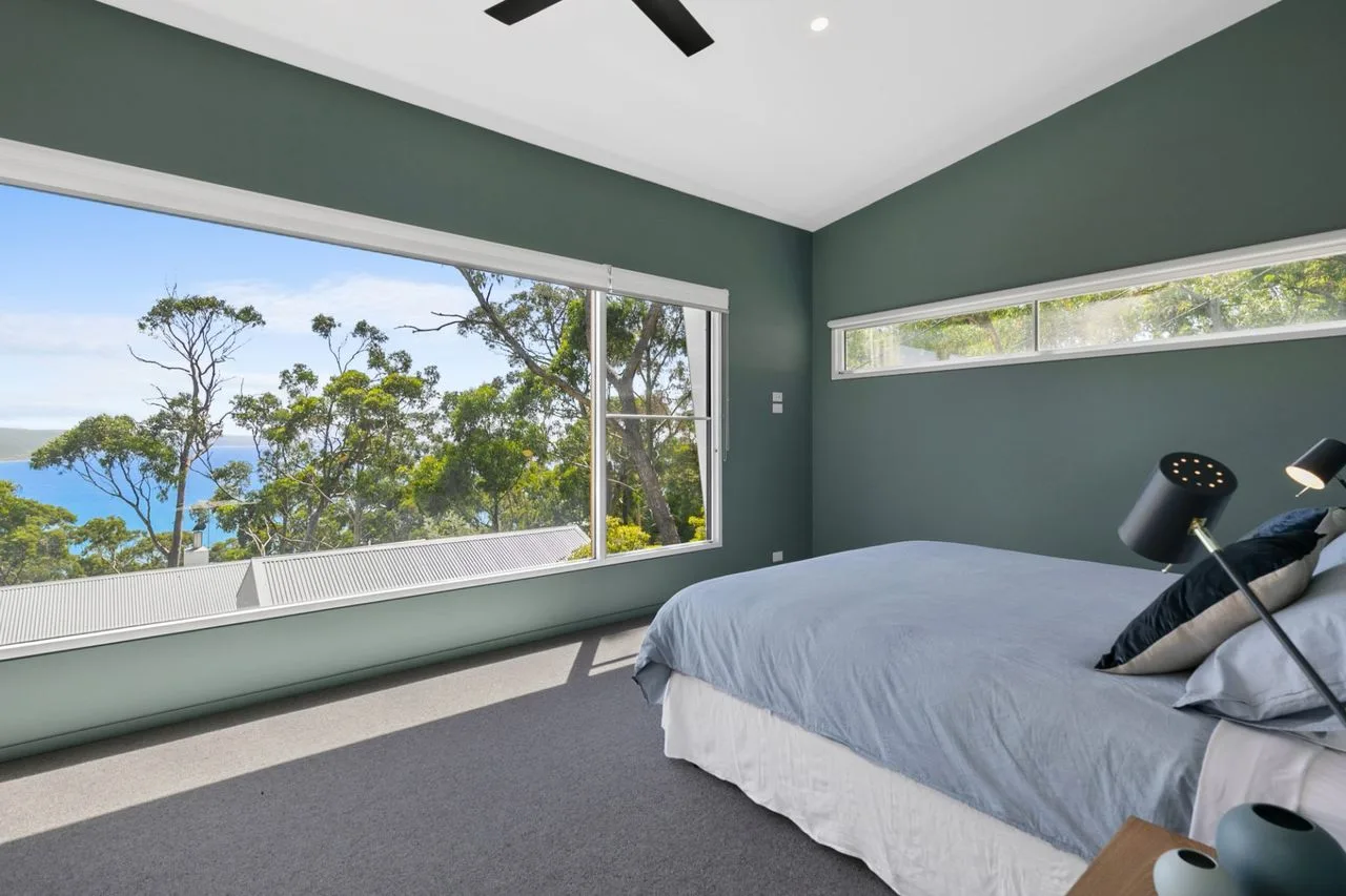 Un dormitorio con una cama junto a grandes ventanas con vistas a árboles y agua.