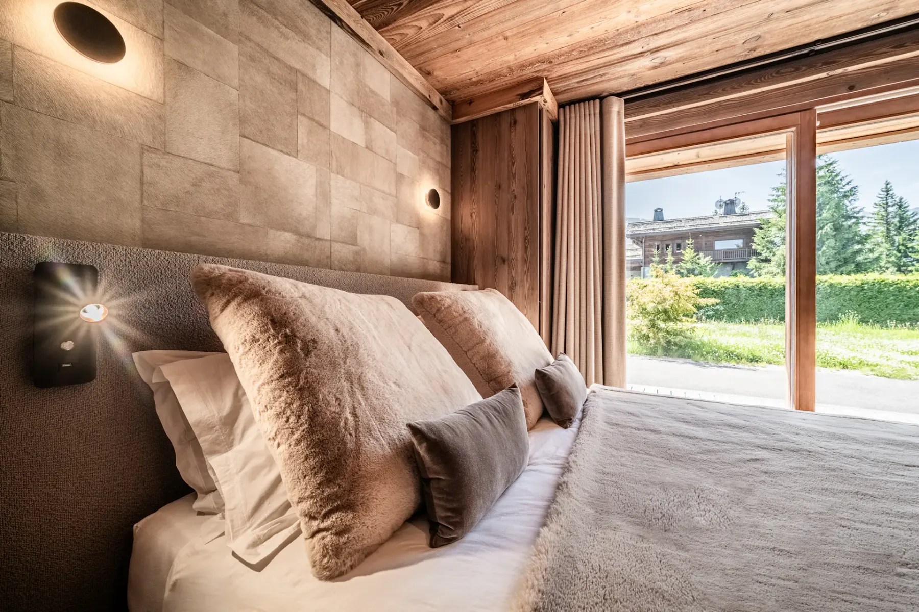 Dormitorio de estilo rústico moderno con paredes y techo revestidos de madera, una cama grande con ropa de cama en tonos neutros, apliques de pared y puertas de vidrio que dan a vistas al jardín.
