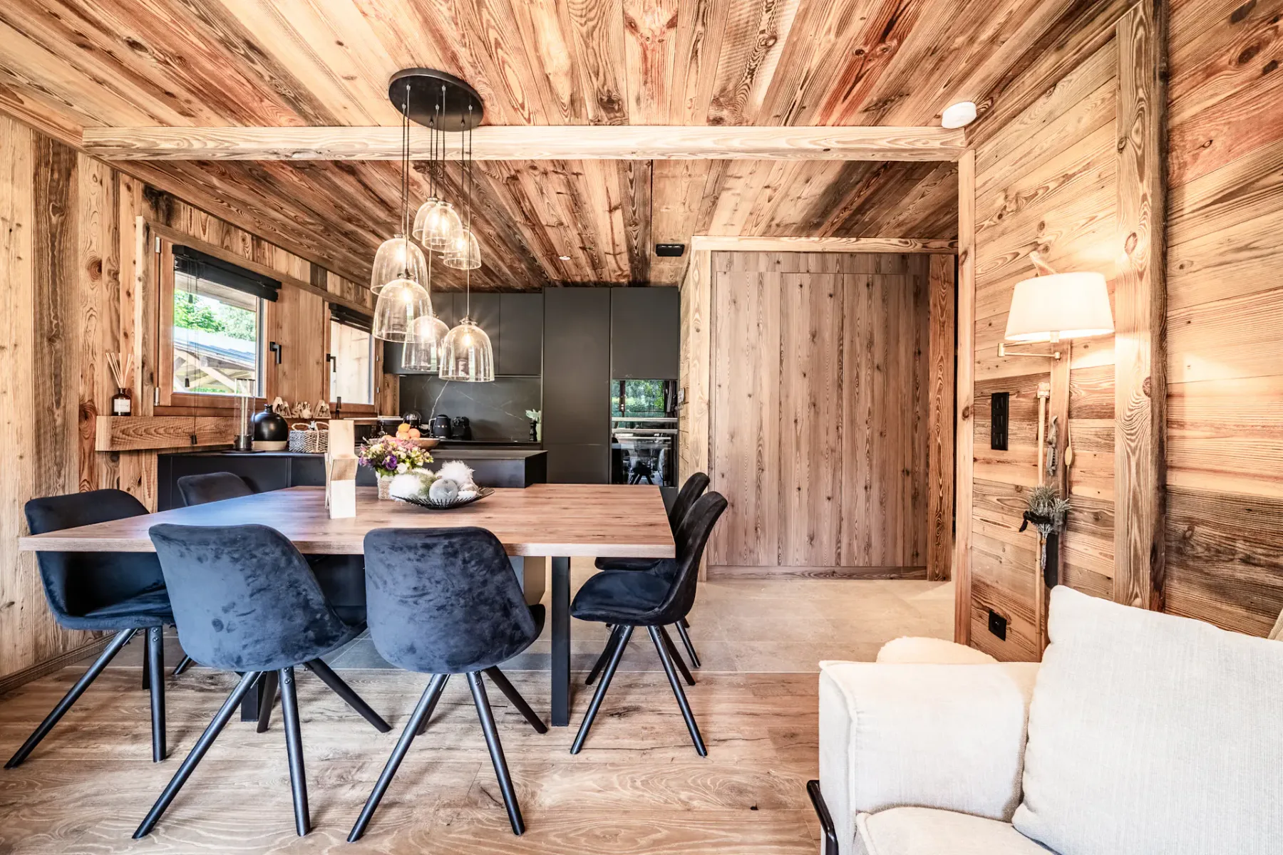Cocina y comedor modernos con armarios negros, paredes y techo de madera, una mesa de madera con sillas negras y lámparas colgantes, en un ambiente interior acogedor y rústico.