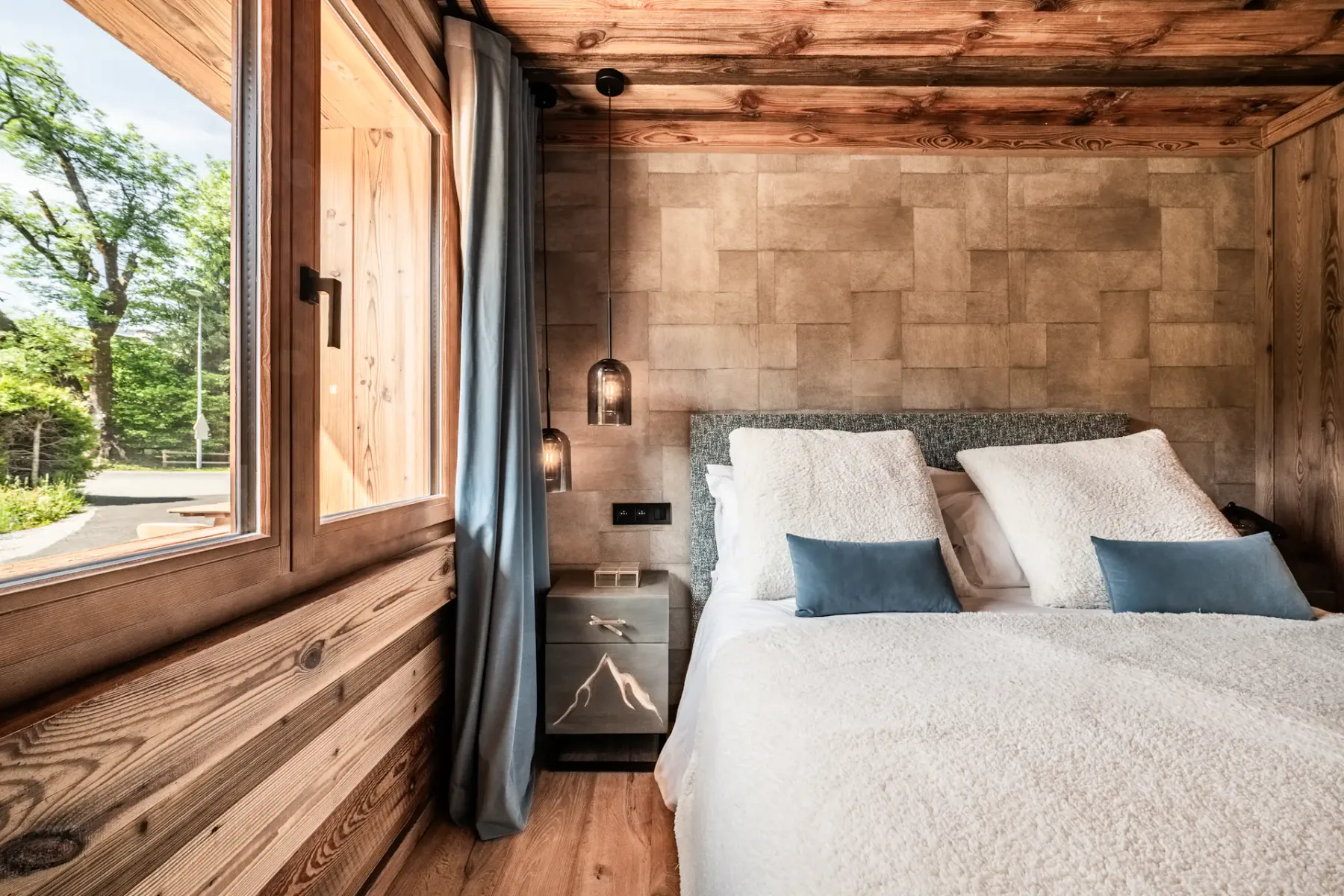 Un dormitorio acogedor con un interior de madera, una cama grande con ropa de cama blanca y cojines decorativos azules, lámparas colgantes y una ventana que deja entrar luz natural.