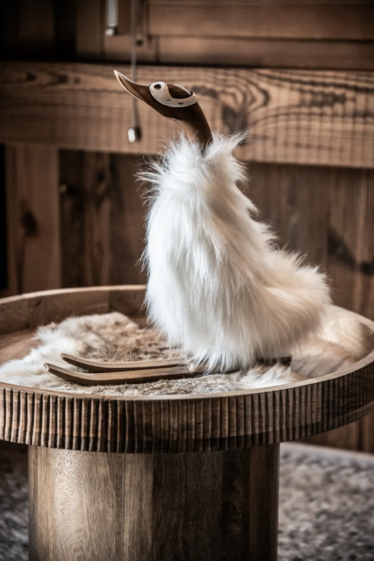 Una figura de pato de madera con el cuerpo de pelo sintético blanco se encuentra sobre una mesa redonda de madera, con un fondo de madera rústica.