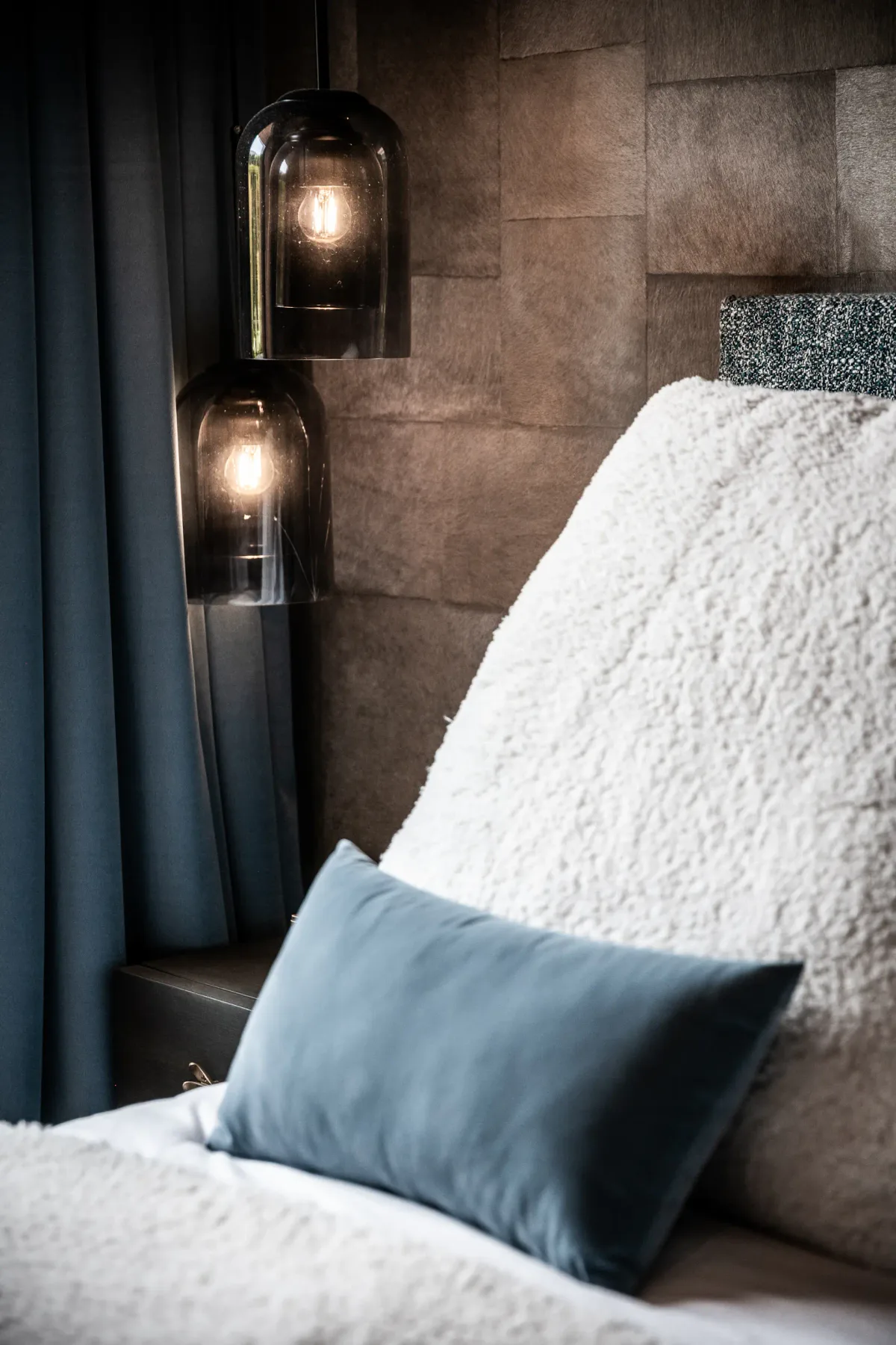 Un dormitorio moderno con una pared texturizada, lámparas colgantes encendidas, una cama con una manta blanca de felpa y una almohada azul.