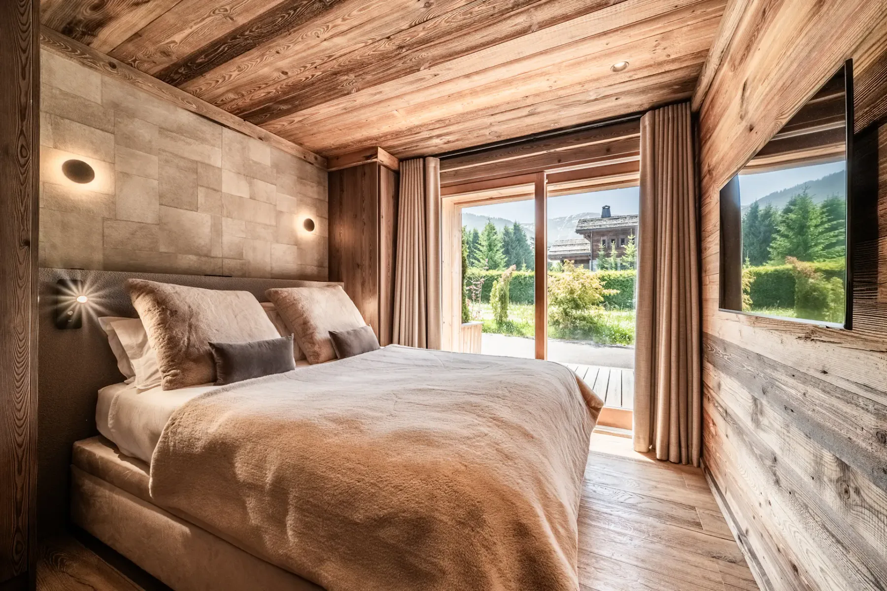 Dormitorio de estilo rústico moderno con paredes y techo revestidos de madera, una cama grande con ropa de cama en tonos neutros, apliques de pared y puertas de vidrio que dan a vistas al jardín.