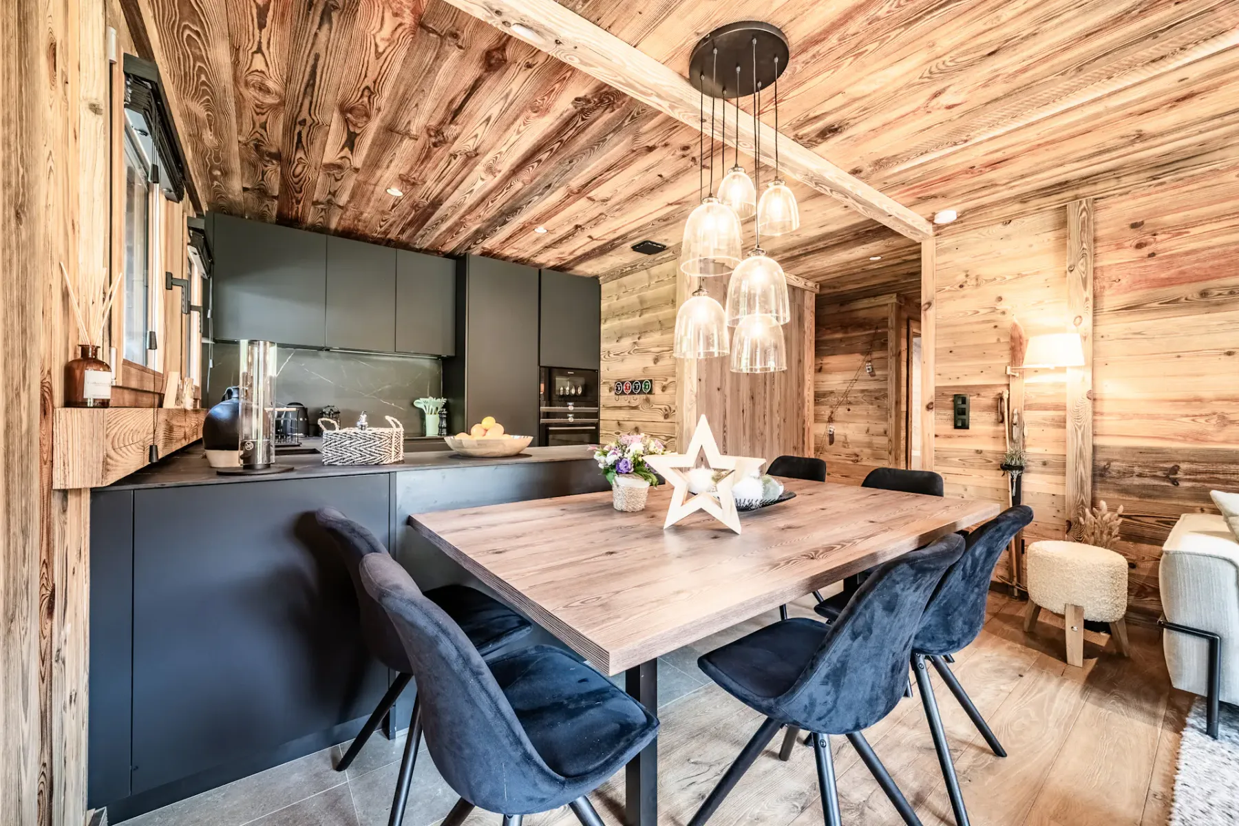 Cocina y comedor modernos con armarios negros, paredes y techo de madera, una mesa de madera con sillas negras y lámparas colgantes, en un ambiente interior acogedor y rústico.