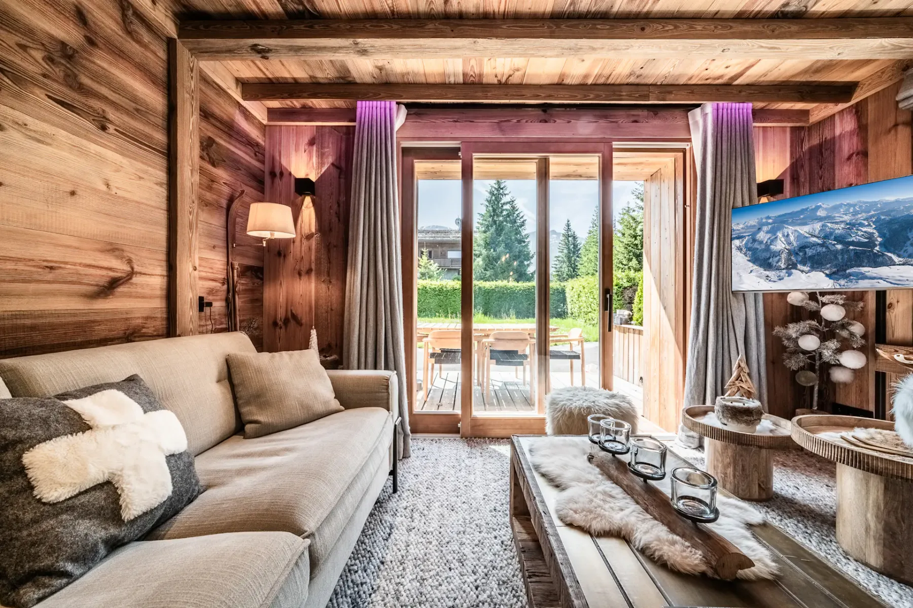 Una acogedora sala de estar con paredes y techo revestidos de madera, un sofá beige, una mesa de centro de madera y grandes puertas de vidrio que dan a un patio soleado con vegetación en el exterior.