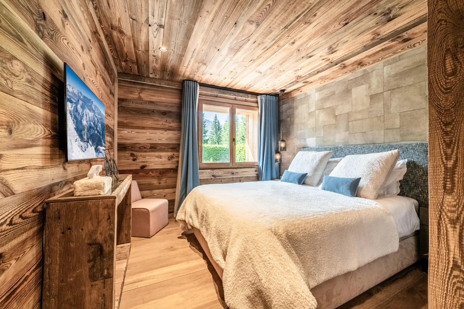 Un dormitorio acogedor con un interior de madera, una cama grande con ropa de cama blanca y cojines decorativos azules, lámparas colgantes y una ventana que deja entrar luz natural.