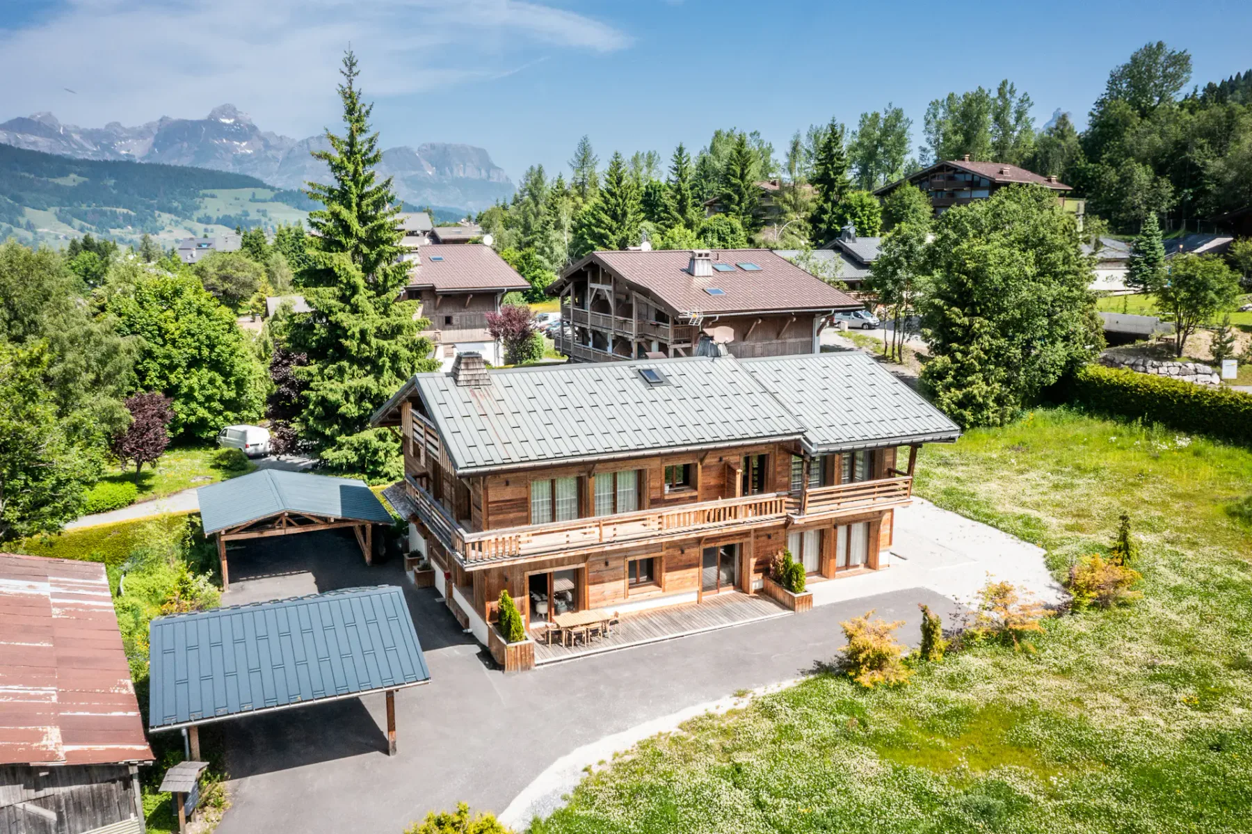 Una casa de madera de estilo chalet con aparcamiento cubierto se encuentra entre árboles y otras casas, en un paisaje montañoso y verde bajo un cielo azul despejado.