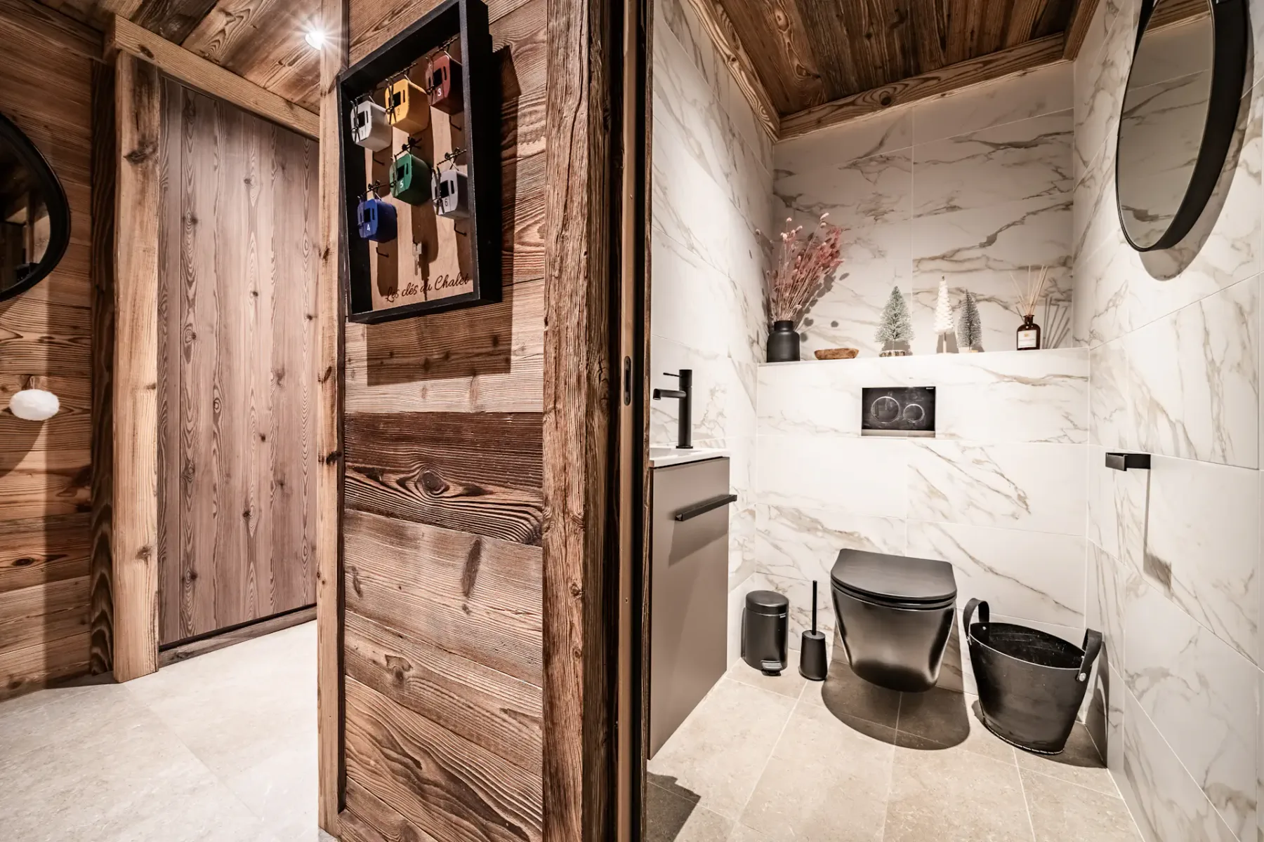 Un baño moderno con paredes de mármol blanco, grifería negra, un espejo redondo, objetos decorativos y una puerta de madera, junto a un pasillo con suelo claro y paredes de madera.