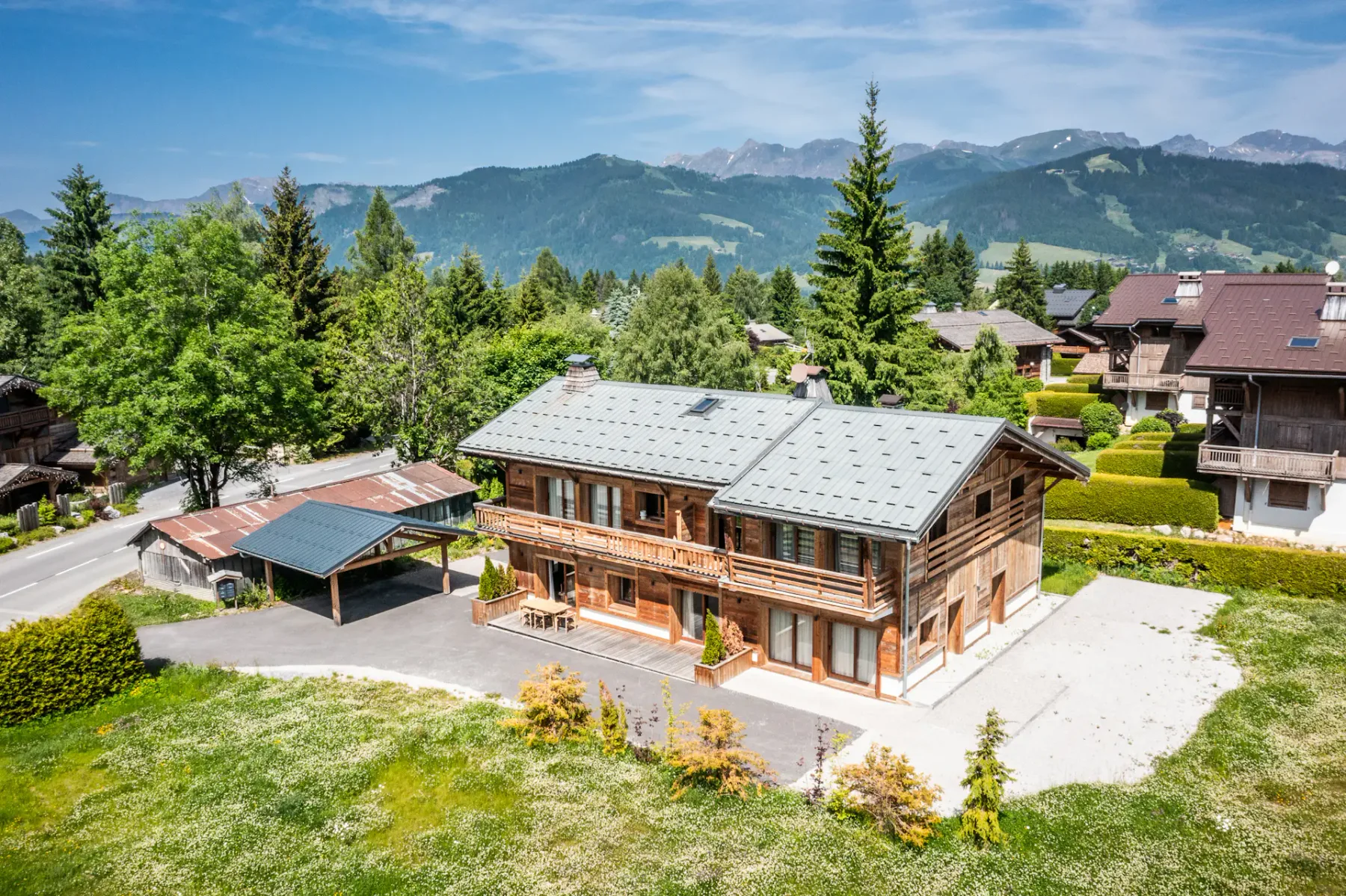 Una casa de madera de estilo chalet con aparcamiento cubierto se encuentra entre árboles y otras casas, en un paisaje montañoso y verde bajo un cielo azul despejado.