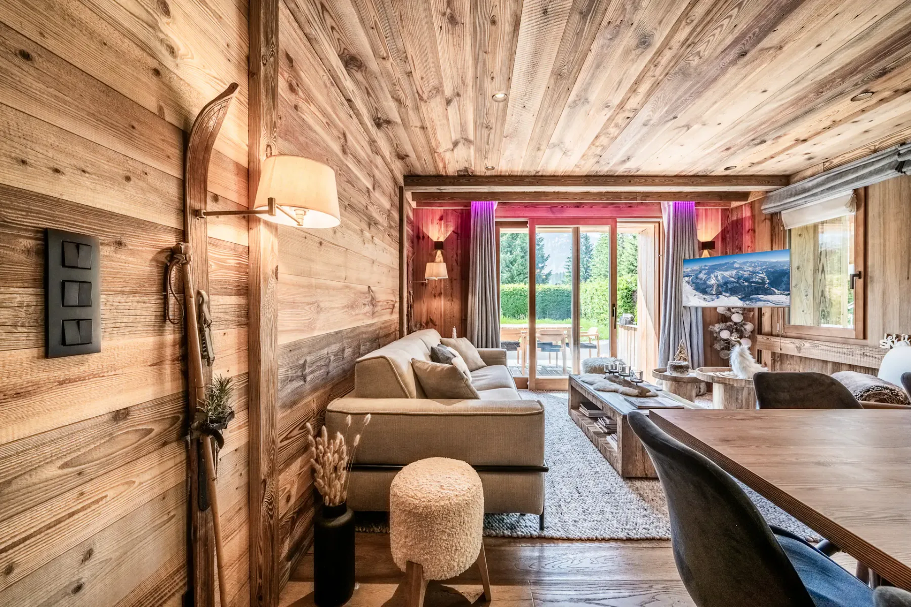 A cozy living room with wood-paneled walls and ceiling, a beige sofa, a wooden coffee table, and large glass doors opening to a sunlit patio with greenery outside.