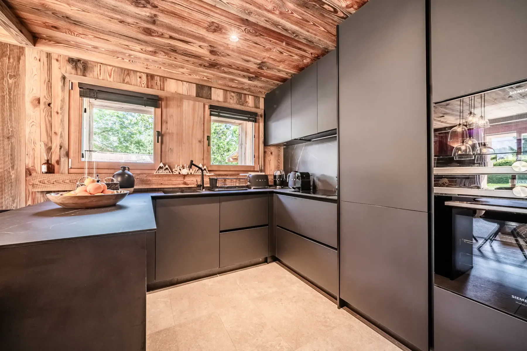 Cocina moderna con armarios oscuros, paredes revestidas de madera y dos ventanas.
