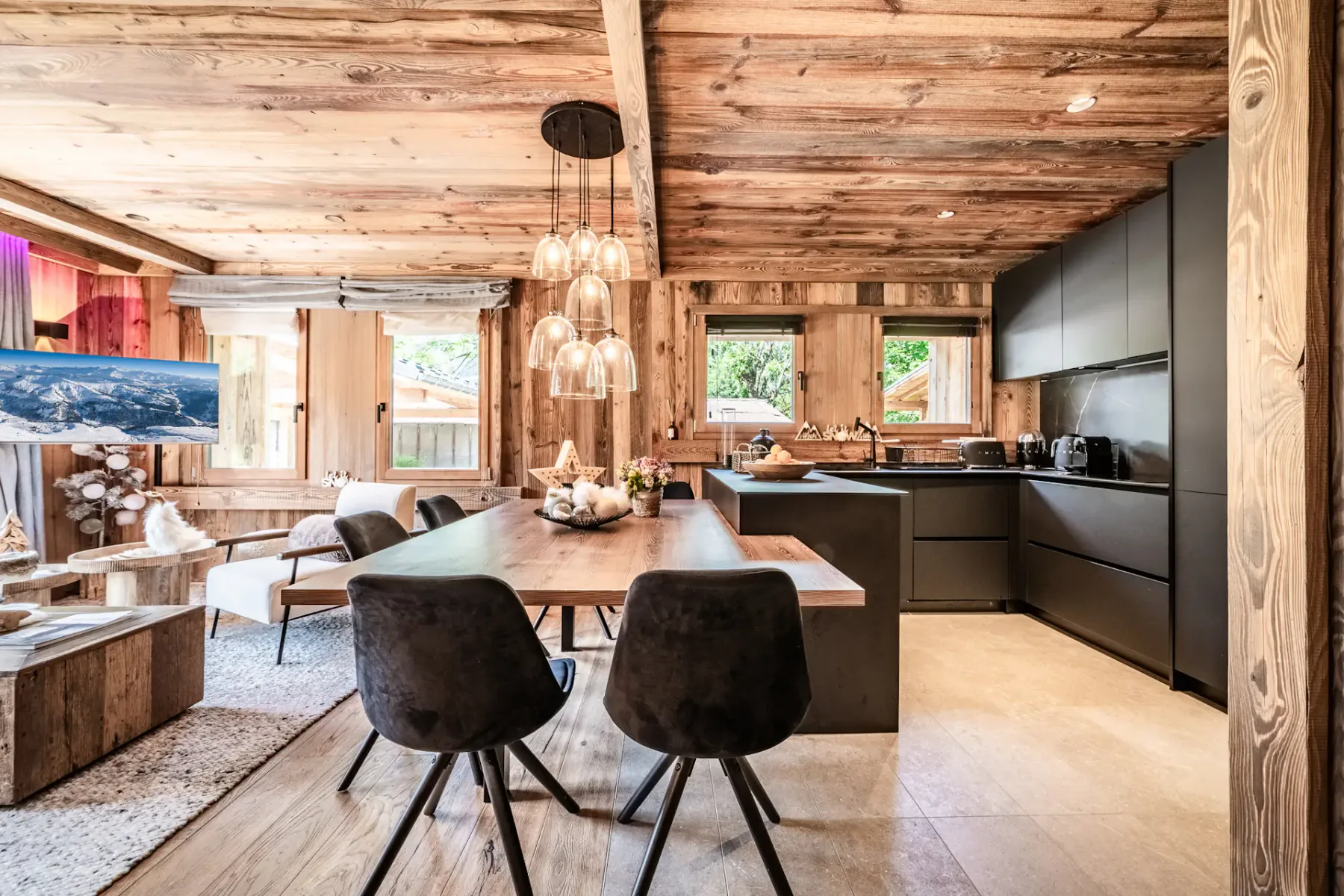 Cocina y comedor modernos con armarios negros, paredes y techo de madera, una mesa de madera con sillas negras y lámparas colgantes, en un ambiente interior acogedor y rústico.