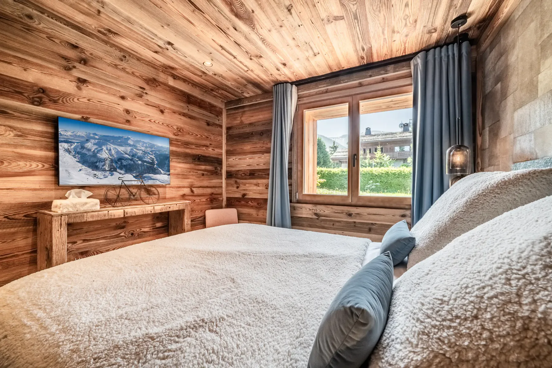 Un dormitorio acogedor con un interior de madera, una cama grande con ropa de cama blanca y cojines decorativos azules, lámparas colgantes y una ventana que deja entrar luz natural.