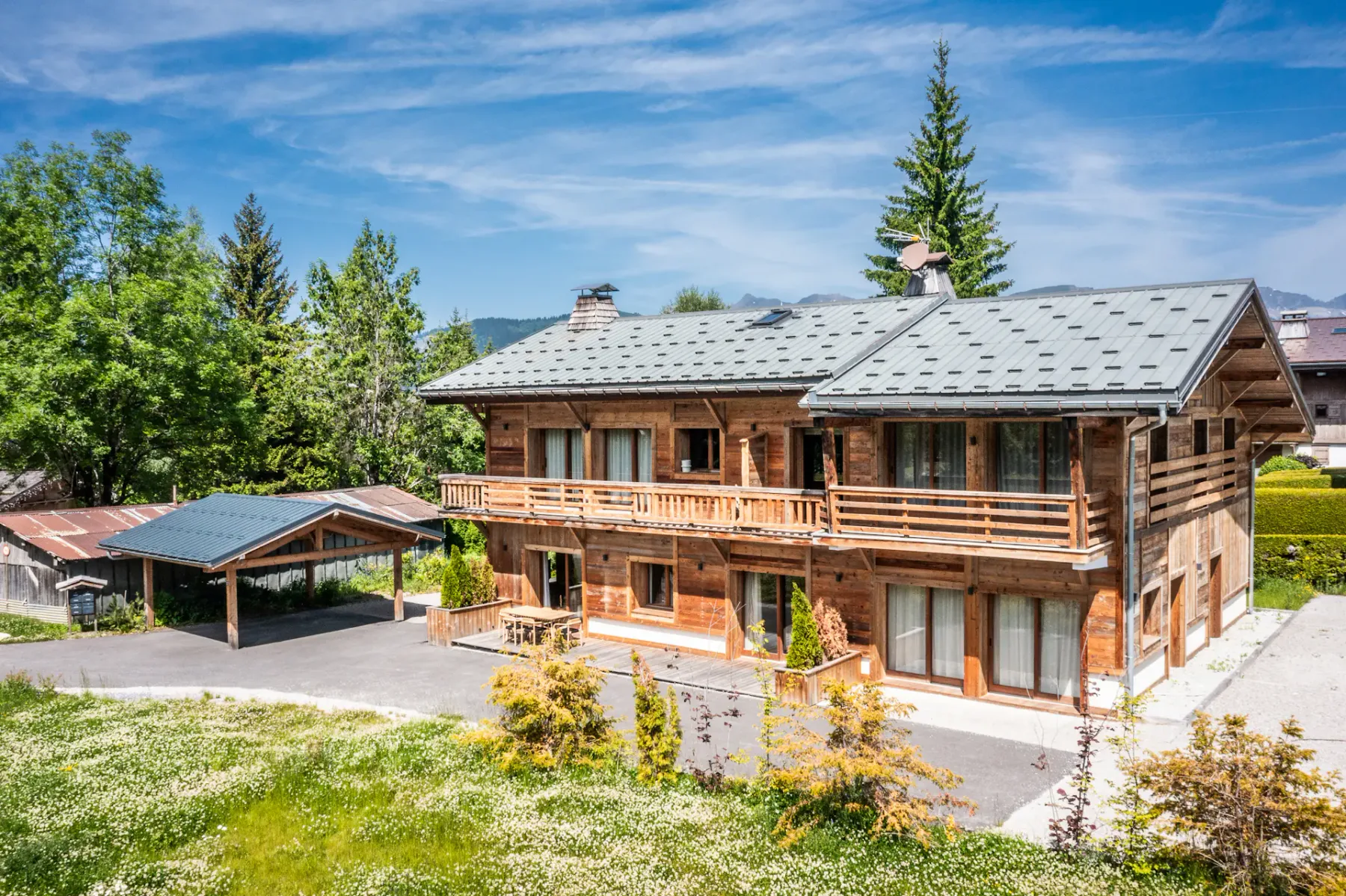 Una casa de madera de estilo chalet con aparcamiento cubierto se encuentra entre árboles y otras casas, en un paisaje montañoso y verde bajo un cielo azul despejado.