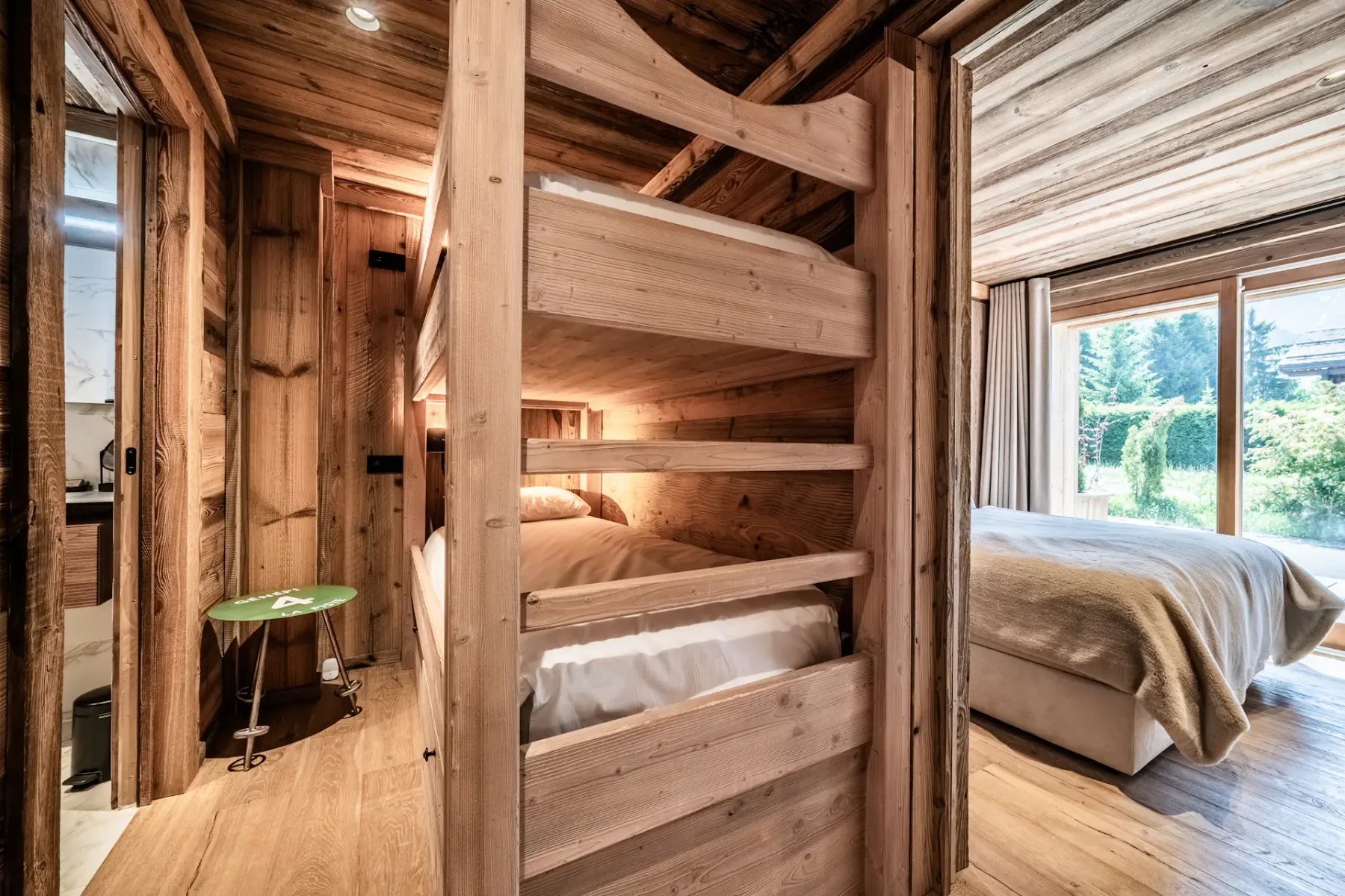 Dormitorio revestido de madera con literas, una pequeña mesa auxiliar verde, una cama doble y una ventana grande con vistas a la vegetación exterior.