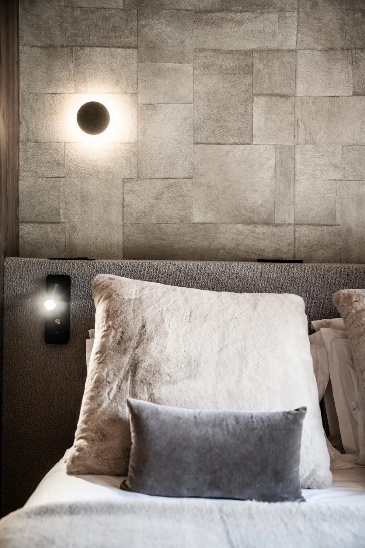 Un dormitorio moderno con una pared gris texturizada, un aplique circular y una cama con grandes almohadas mullidas y un pequeño cojín gris.