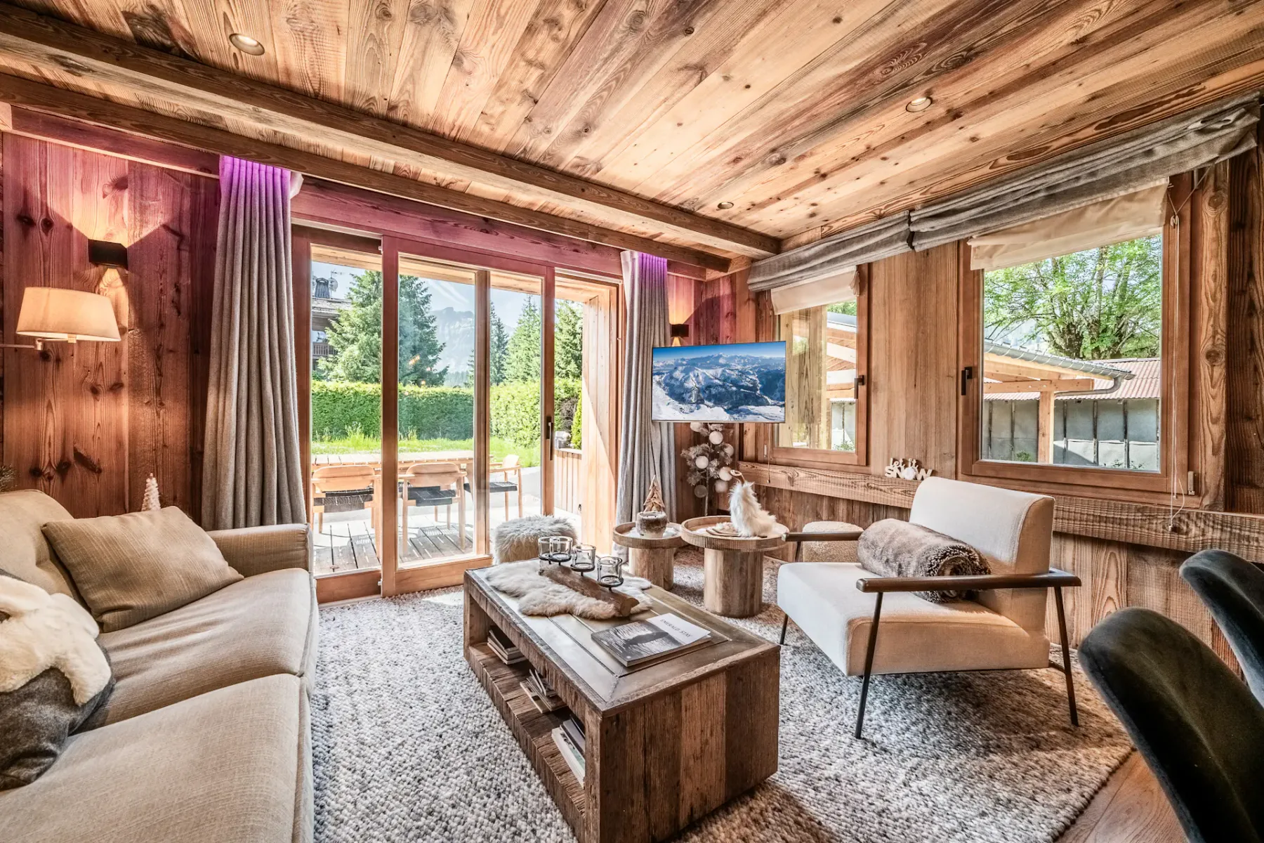 A cozy living room with wood-paneled walls and ceiling, a beige sofa, a wooden coffee table, and large glass doors opening to a sunlit patio with greenery outside.