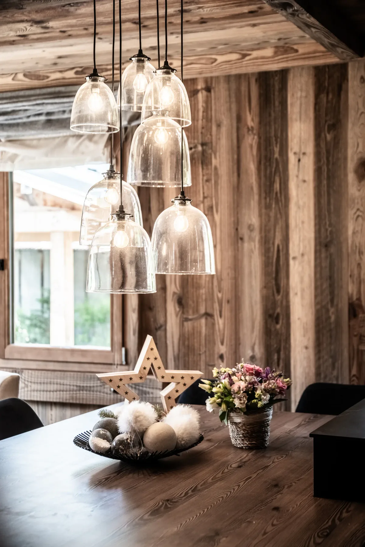 Una mesa de comedor de madera con objetos decorativos, entre ellos una estrella, un cuenco con adornos y un arreglo floral, situada bajo lámparas colgantes de vidrio en una estancia rústica con paredes revestidas de madera.