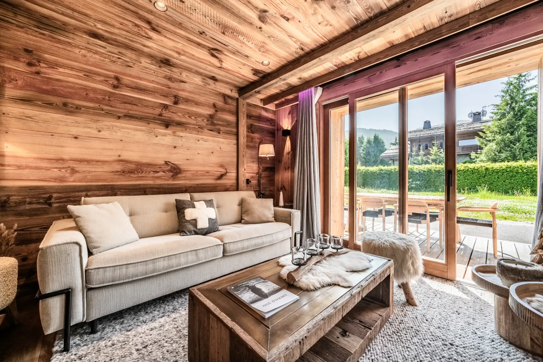 Una acogedora sala de estar con paredes y techo revestidos de madera, un sofá beige, una mesa de centro de madera y grandes puertas de vidrio que dan a un patio soleado con vegetación en el exterior.
