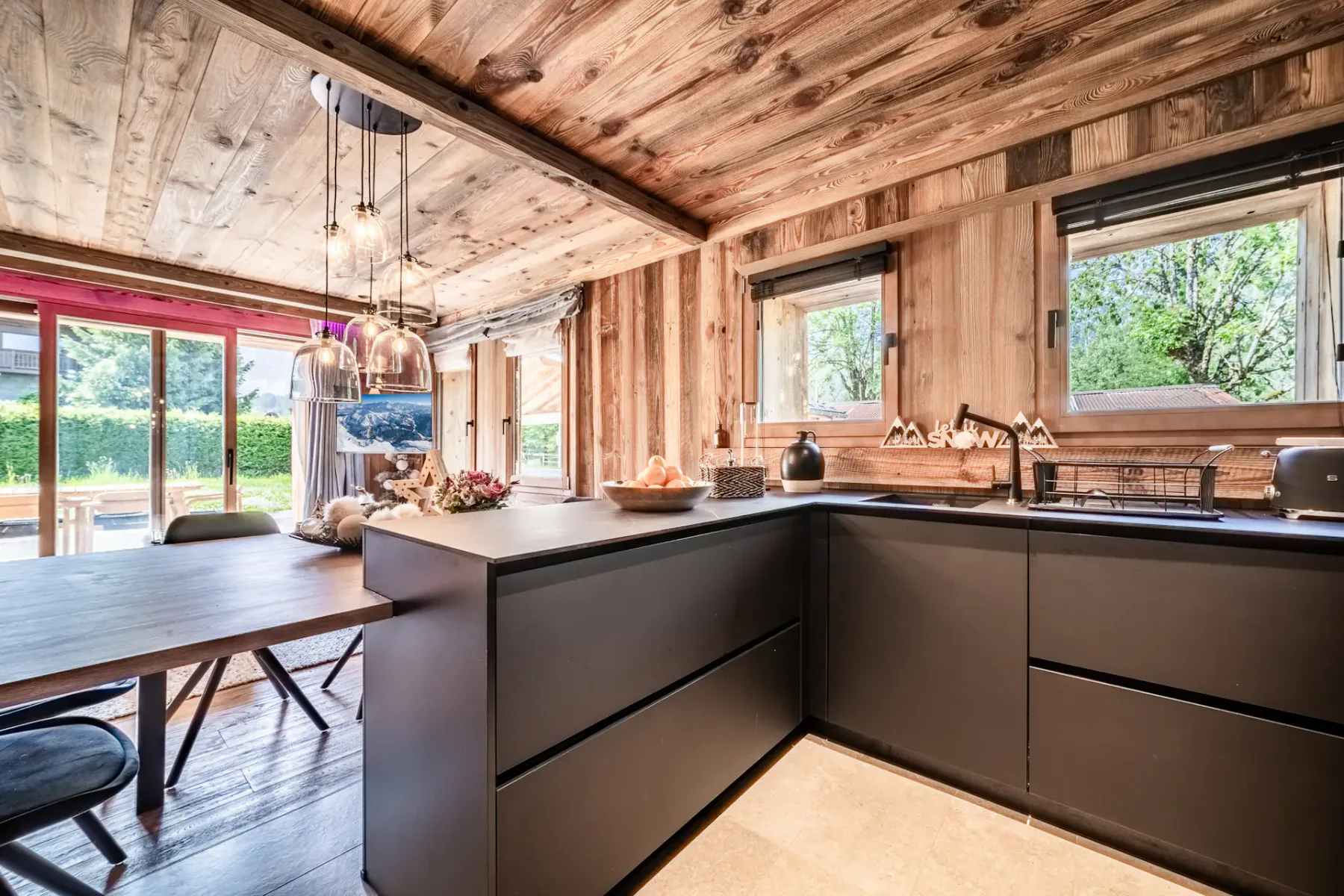 Cocina y comedor modernos con armarios negros, paredes y techo de madera, una mesa de madera con sillas negras y lámparas colgantes, en un ambiente interior acogedor y rústico.