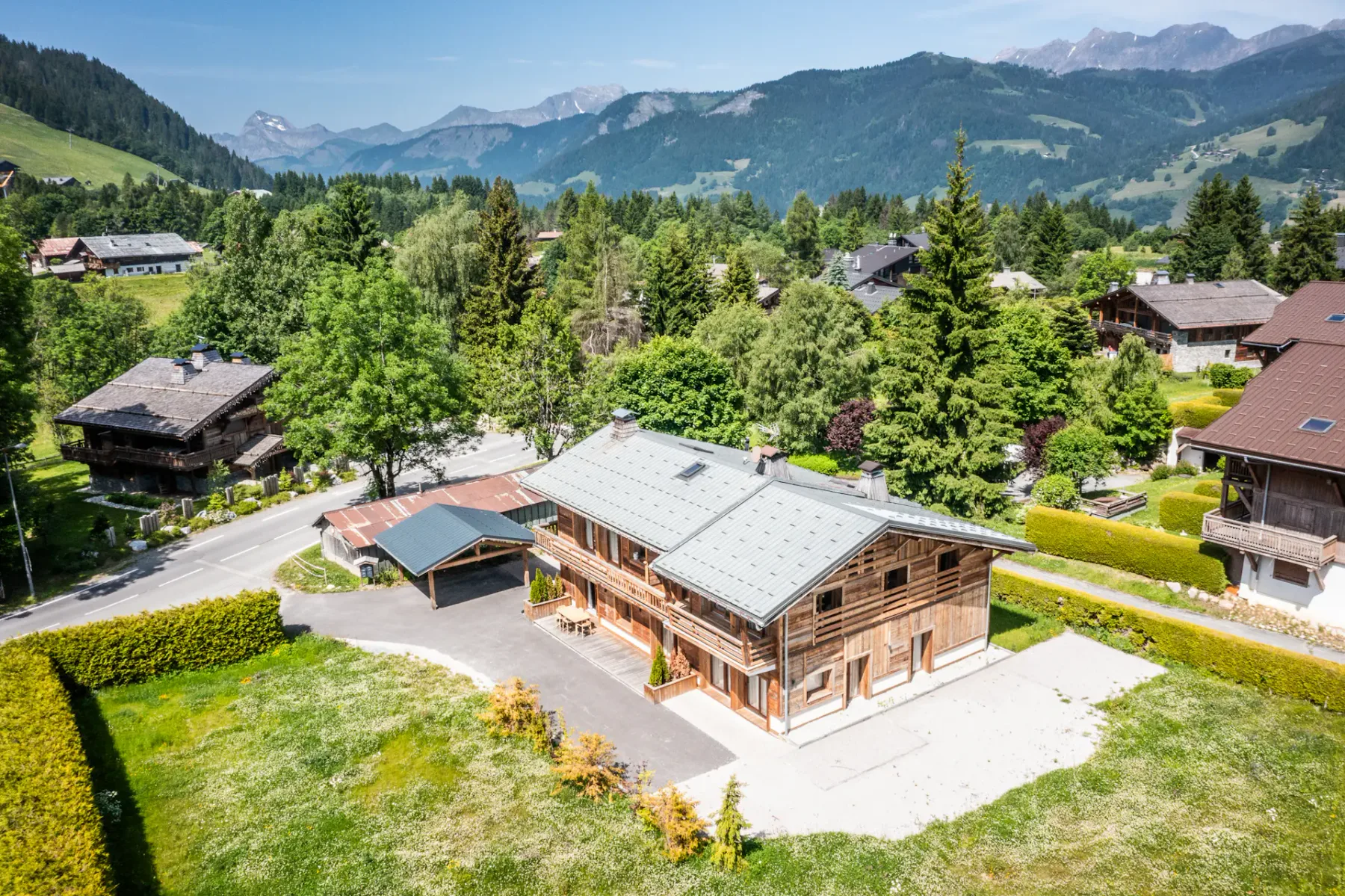 Un edificio de estilo chalet de madera está entre árboles y casas vecinas, con montañas verdes al fondo.