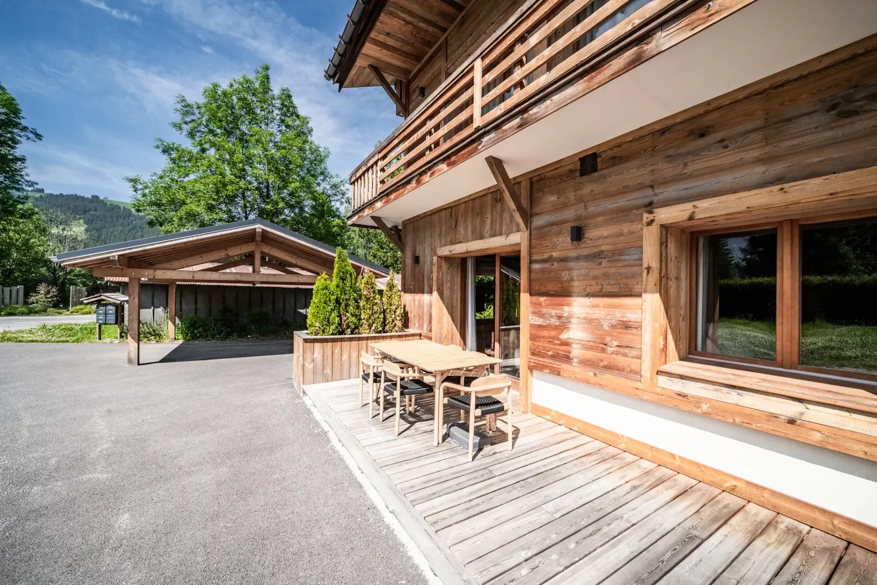 Un chalet de madera con una amplia terraza, mesa de comedor exterior, sillas, grandes ventanales y un balcón, situado junto a vegetación y un acceso pavimentado para vehículos.