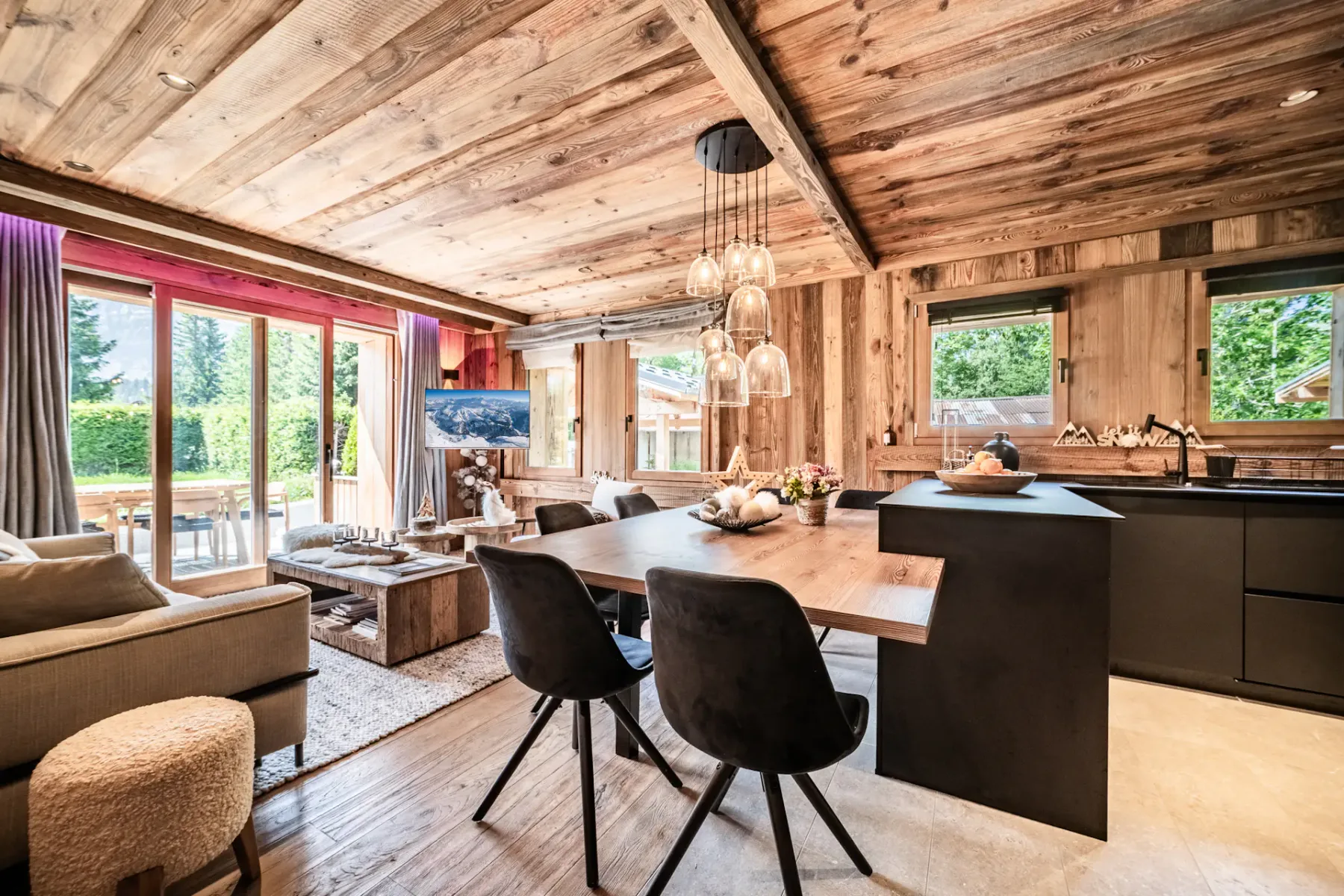 Cocina y comedor modernos con armarios negros, paredes y techo de madera, una mesa de madera con sillas negras y lámparas colgantes, en un ambiente interior acogedor y rústico.