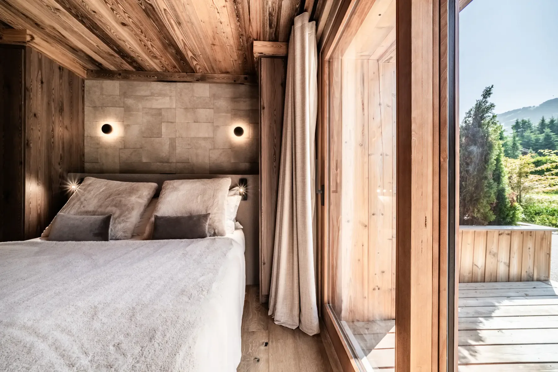 Dormitorio de estilo rústico moderno con paredes y techo revestidos de madera, una cama grande con ropa de cama en tonos neutros, apliques de pared y puertas de vidrio que dan a vistas al jardín.
