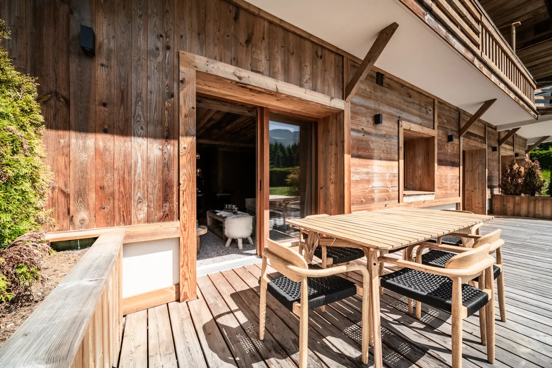 Un chalet de madera con una amplia terraza de madera, una mesa de comedor exterior, sillas, grandes ventanas y un balcón, situado junto a vegetación y una entrada pavimentada para vehículos.