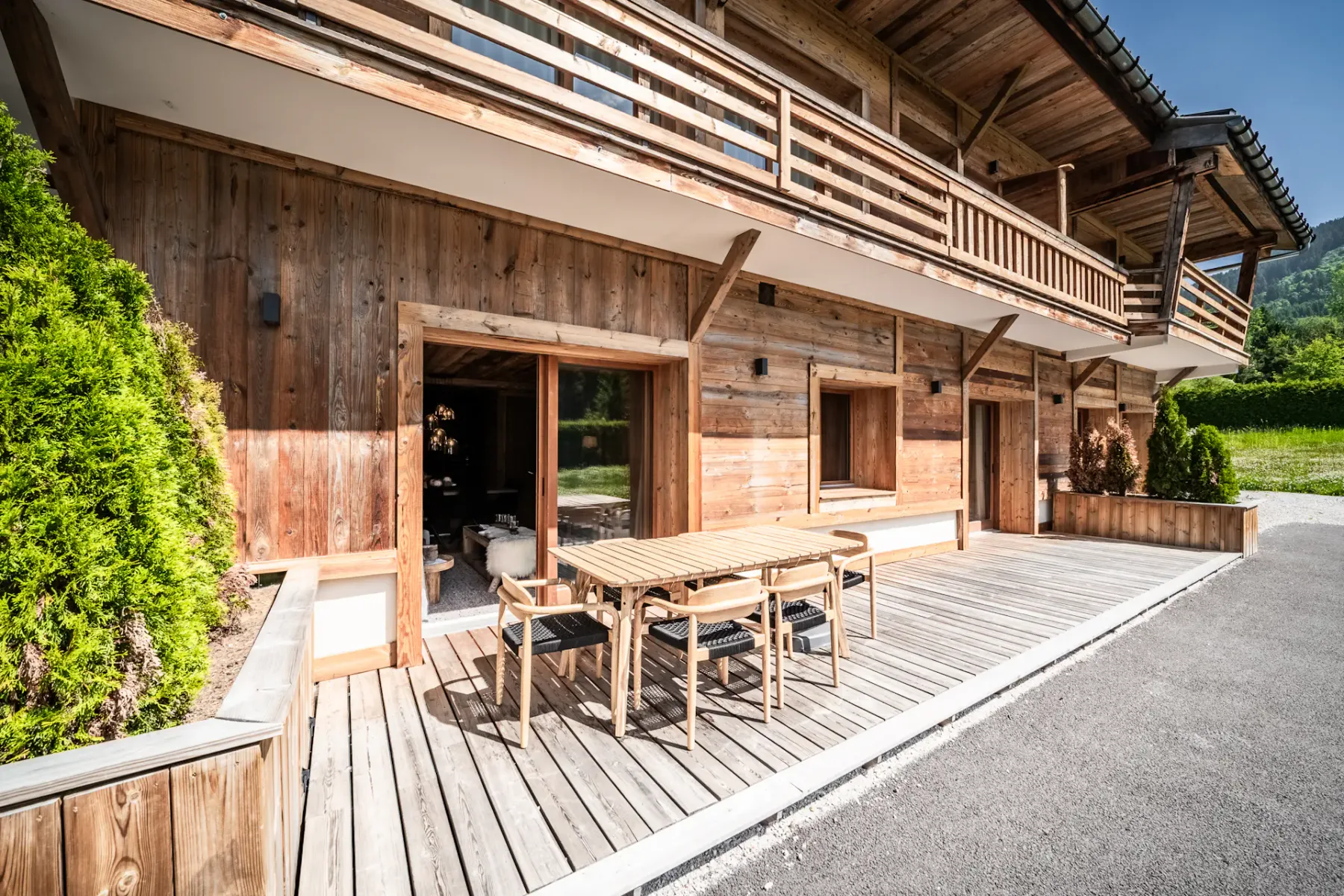 Un chalet de madera con una amplia terraza de madera, una mesa de comedor exterior, sillas, grandes ventanas y un balcón, situado junto a vegetación y una entrada pavimentada para vehículos.