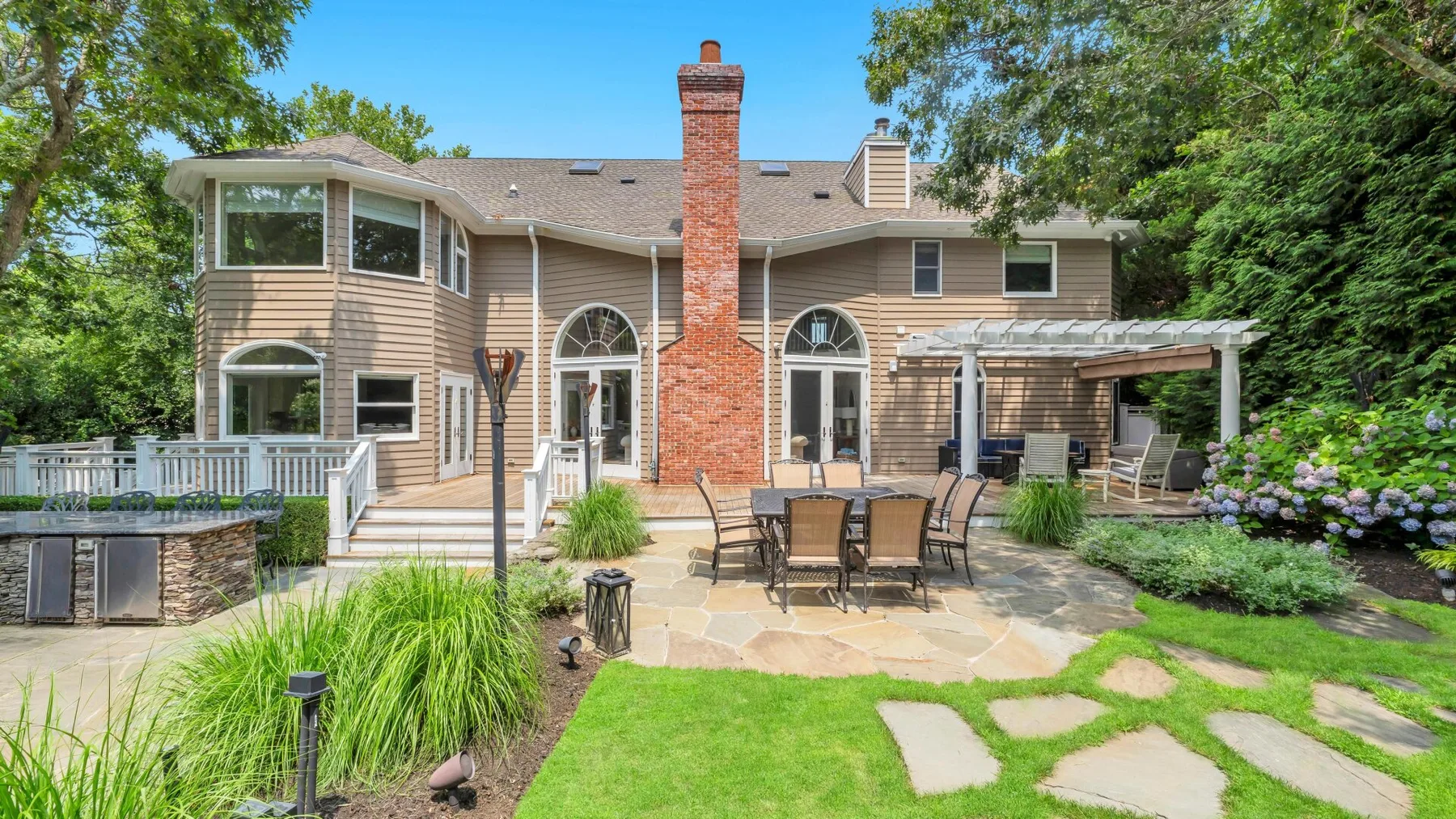A two-story house opens onto a stone patio with outdoor seating and a covered pergola.