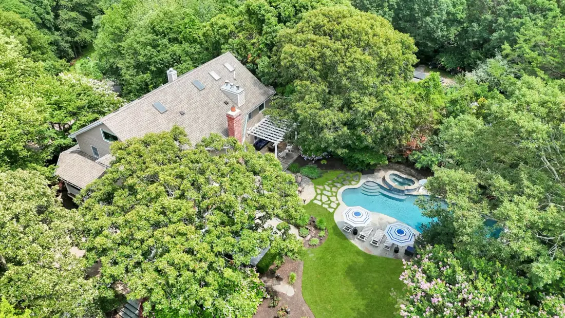 An aerial view shows a house, swimming pool, and green garden surrounded by trees.