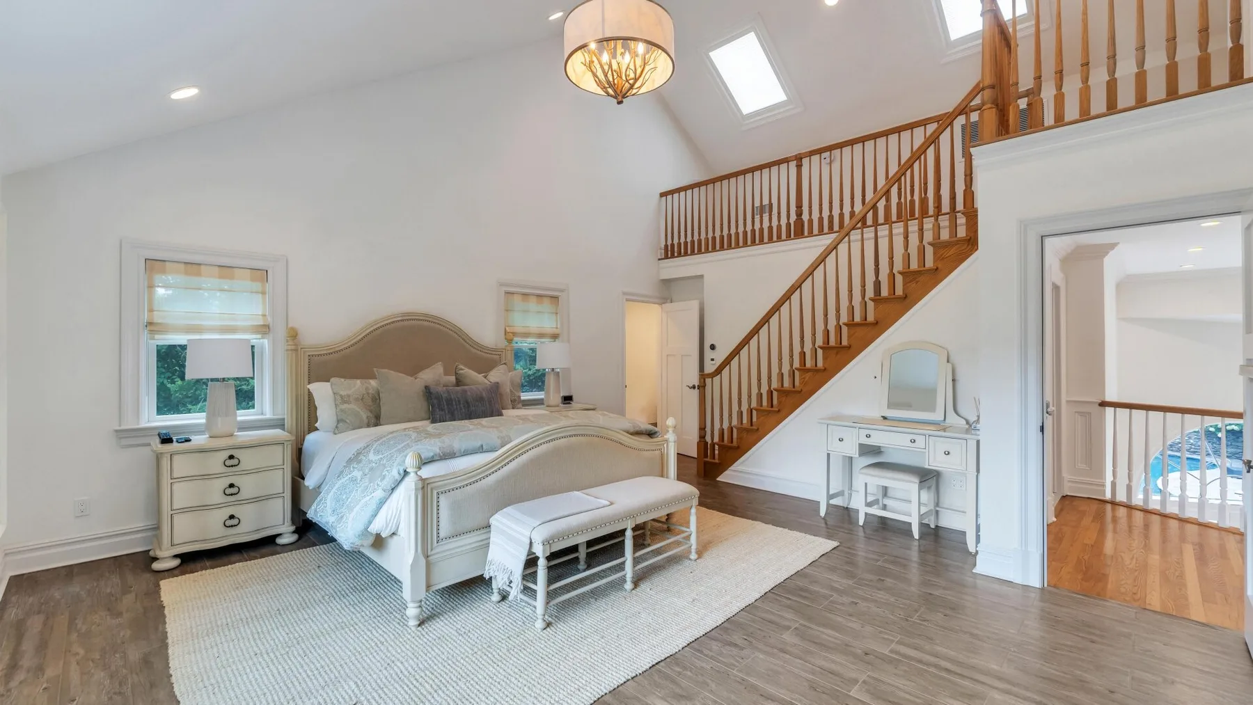 A bright bedroom with a bed, a vanity, and a wooden staircase.