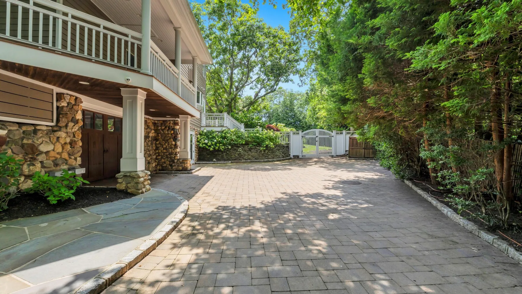 A paved driveway beside a two-story house, with trees and greenery along the edge.
