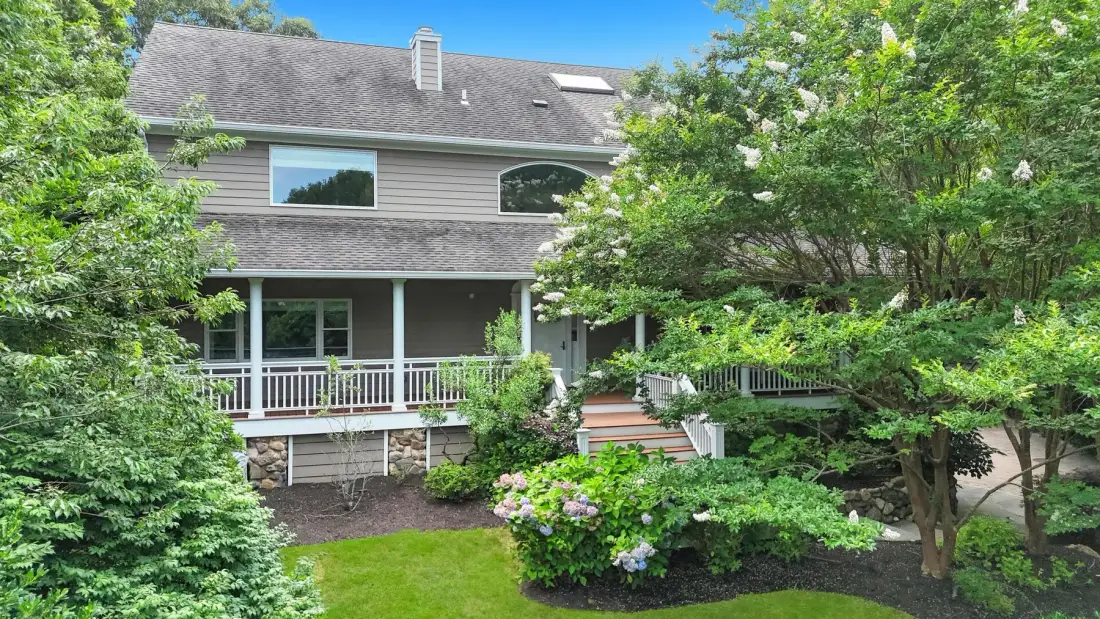 A house with a raised deck and steps sits among leafy trees and flowering shrubs.