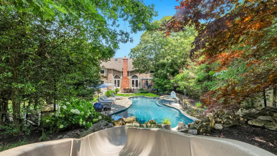 A backyard swimming pool is surrounded by trees and landscaped greenery.