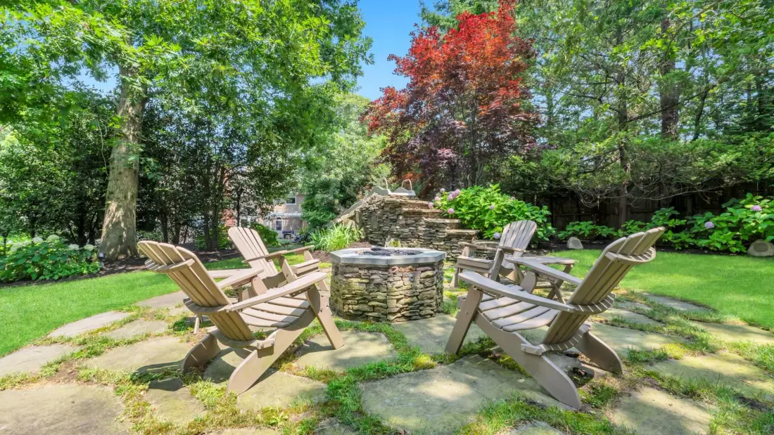 An outdoor seating area surrounds a round stone fire pit among trees and garden greenery.