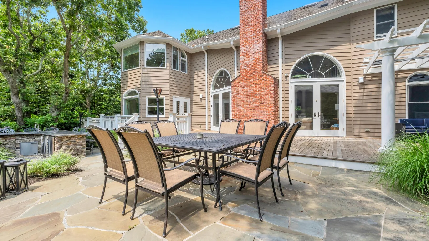 A stone patio with outdoor seating sits beside a tan house and brick chimney.