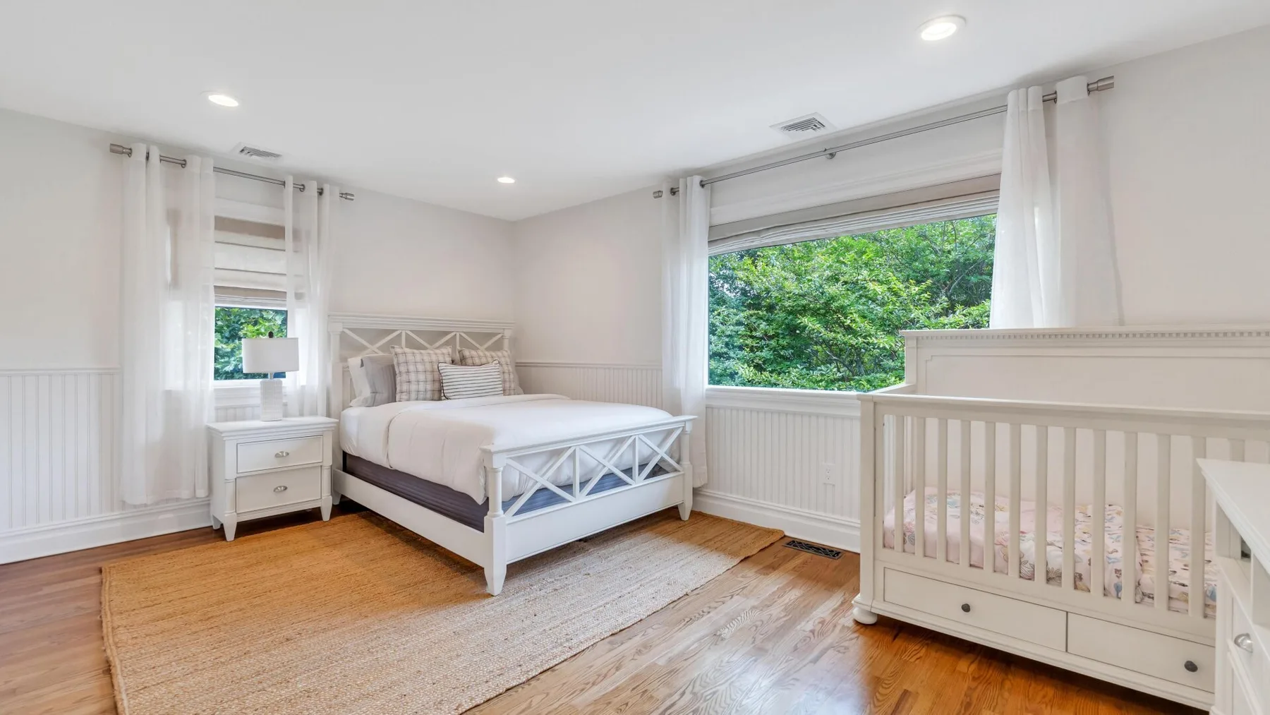 A bright bedroom with a white bed and a crib beside the window.