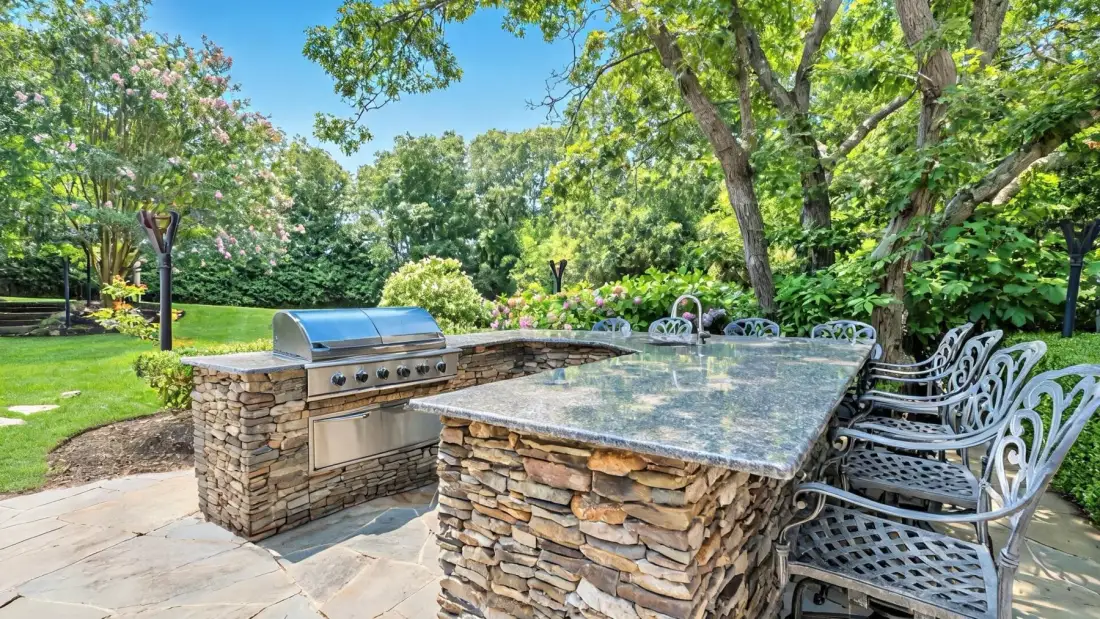 An outdoor stone kitchen and dining area sit in a leafy garden.
