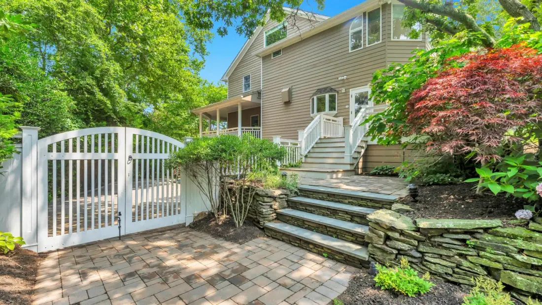 A house exterior with a white gate, stone steps, and surrounding greenery.
