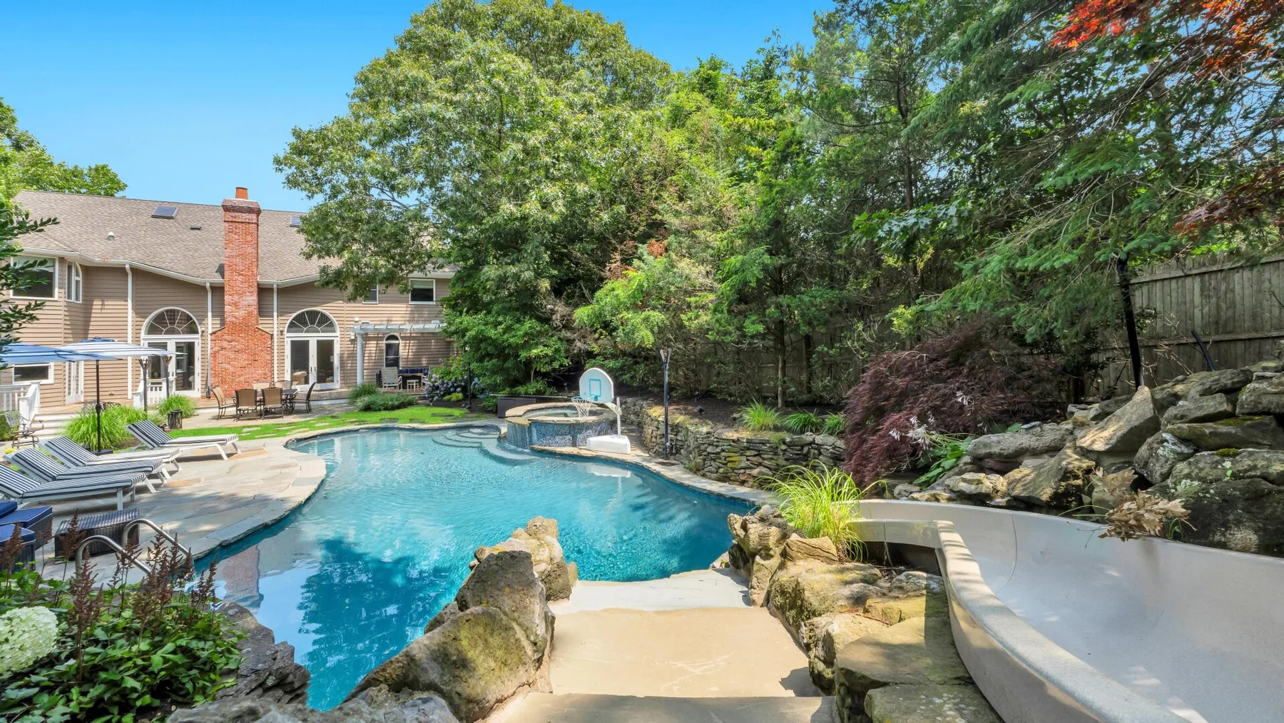 A backyard pool beside a house, surrounded by trees and landscaped rocks.