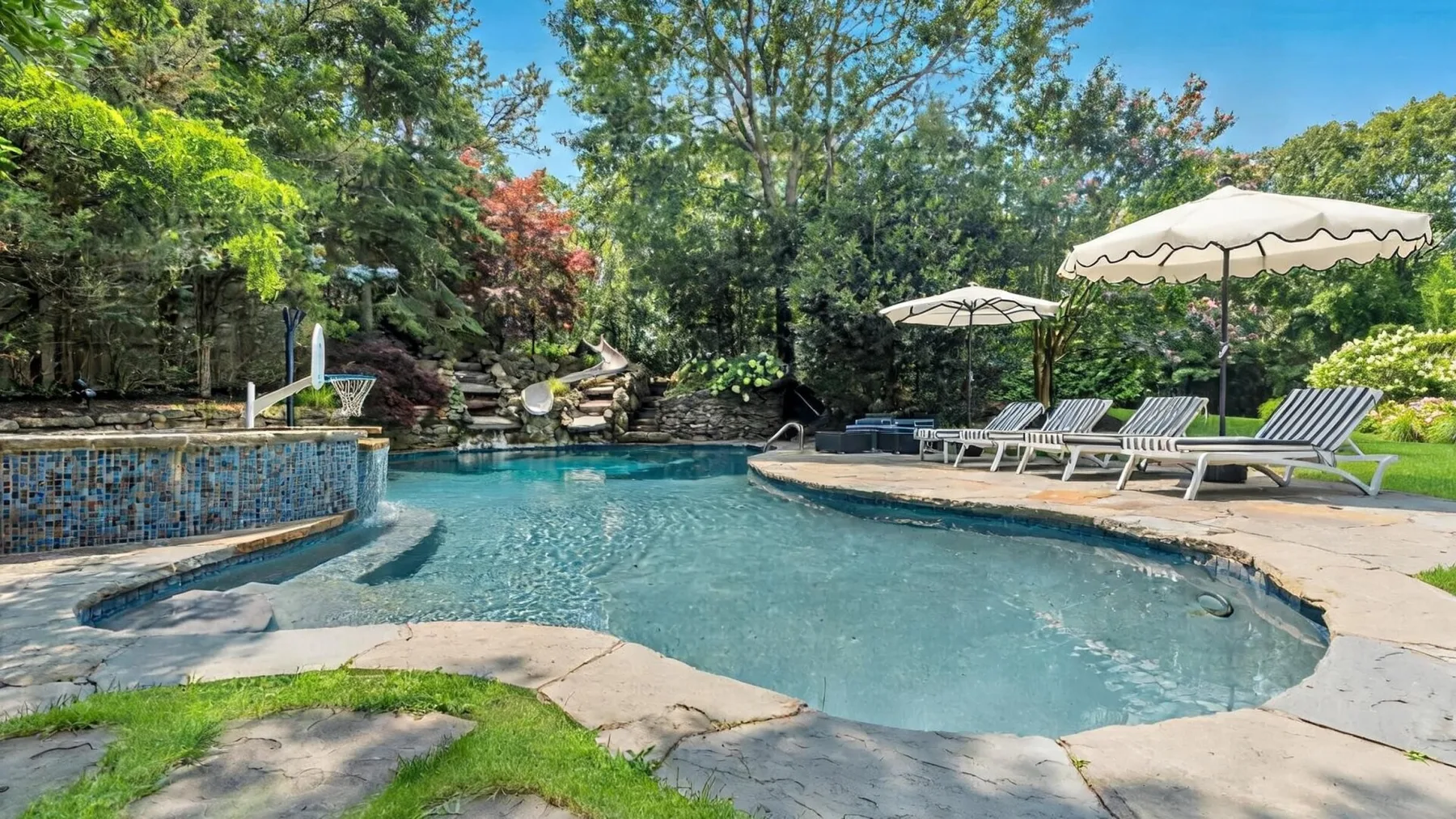 An outdoor swimming pool surrounded by trees, with lounge chairs and umbrellas along the edge.