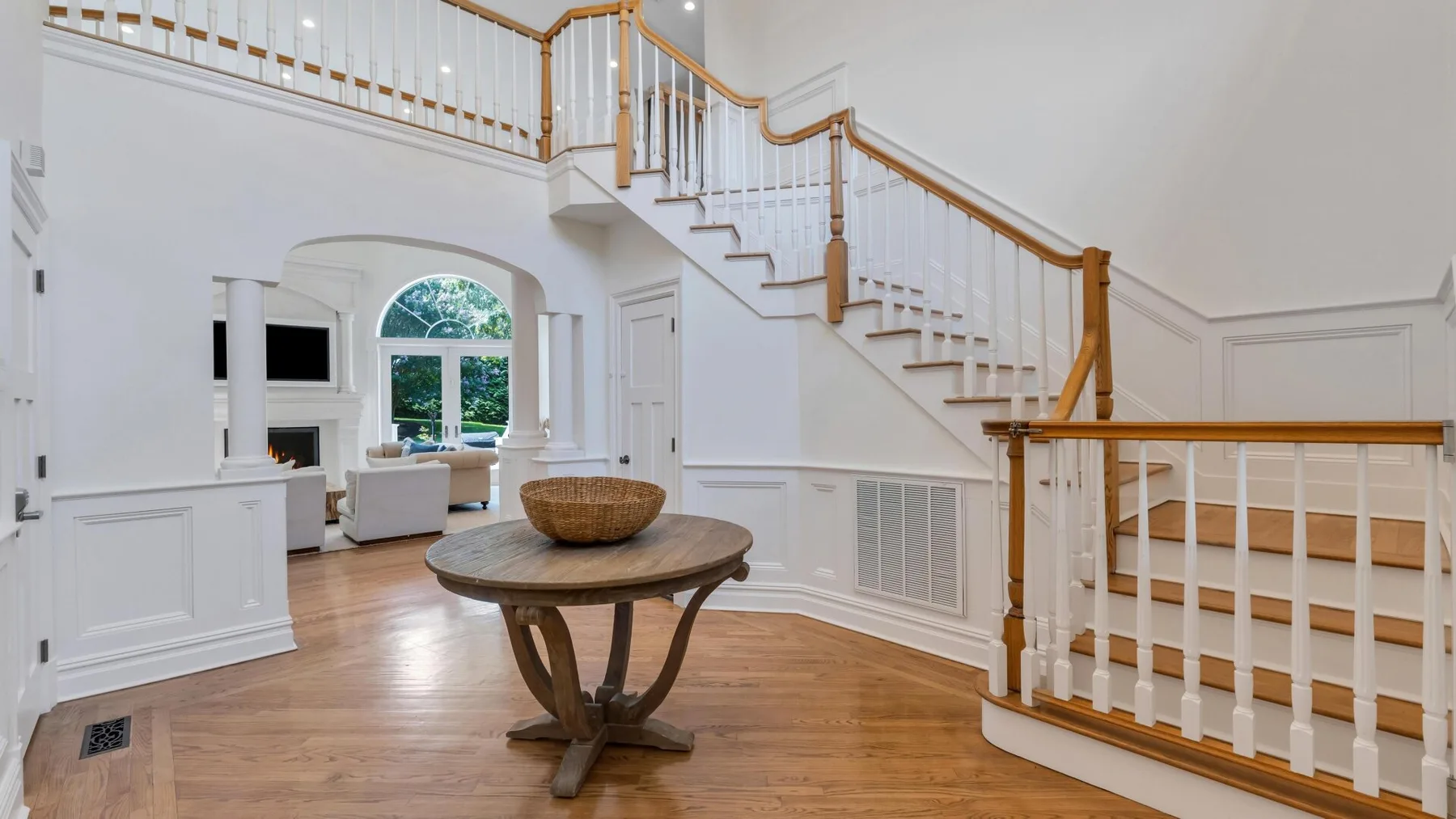 A bright living area beneath a staircase, with a round wooden table in the foreground.