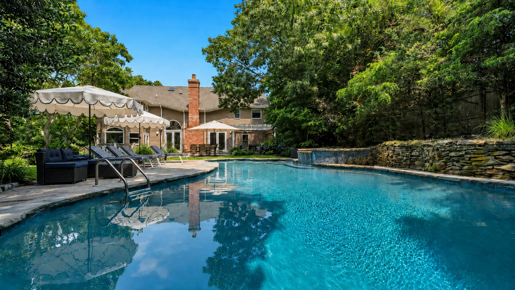 A backyard swimming pool sits beside a house and shaded seating area.