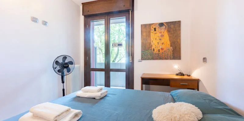 A bedroom with a bed, folded towels, and a desk beneath a framed painting.