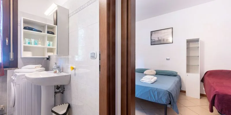 A bathroom with a sink and shelves opens onto a bedroom with beds.
