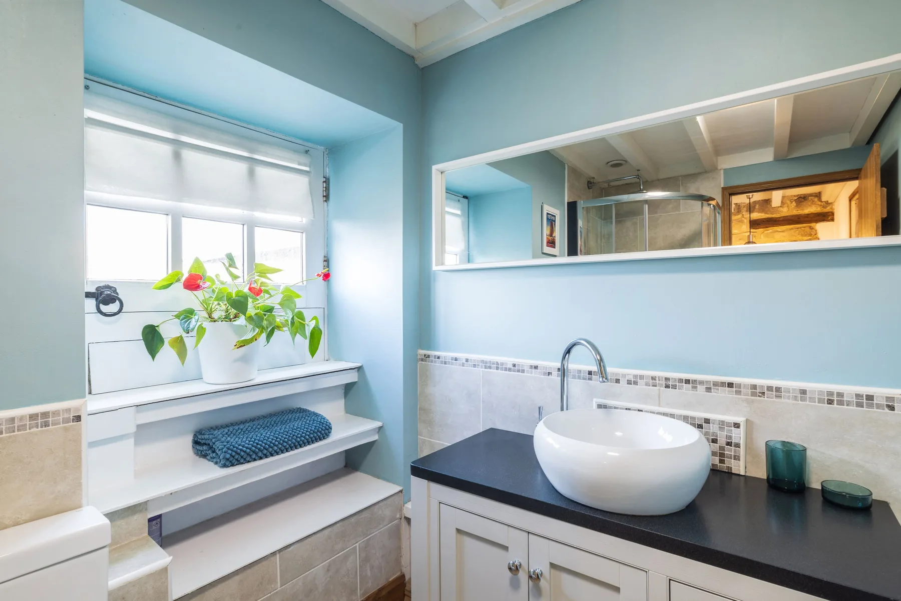 Un tocador de baño con lavabo sobre encimera está junto a una ventana con plantas.