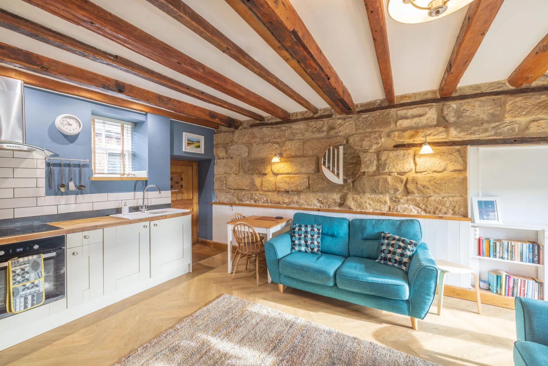 A living room with a teal sofa, a kitchen area, and exposed ceiling beams.